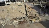 A Syrian rides a bicycle past a collapsed building on which graffiti in Arabic and English reads 