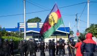 A pro-independence supporter holds a Kanak and Socialist National Liberation Front (FLNKS) flag in front of police officers outside the headquarters of the Union Caledonienne (UC), after police intervened in the UC offices this morning, where a press conference of the Cellule de Coordination des Actions de Terrain (CCAT) was due to be held, in Noumea, on June 19, 2024. Photo by Delphine Mayeur / AFP.
