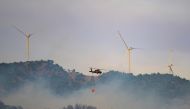 This photograph shows a helicopter carrying water to fight a forest fire, with windmills in the background in Turkey's western province of Izmir on August 17, 2024. (Photo by Yasin AKGUL / AFP)