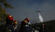Firefighters gather on a field near the area where an airplane drops water over a wildfire that broke up in Pikermi, some 30 Kms east of Athens on July 3, 2025. Photo by Aris MESSINIS / AFP.
