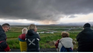 Tourists standing next to a memorial in Keflavik, Iceland on July 16, 2025, observe from a distance smoke and lava emanating from a volcano near Grindavik on the Icelandic peninsula of Reykjanes following its eruption. Photo by Halldor KOLBEINS / AFP