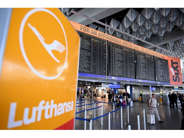 The logo of German airline Lufthansa is seen in the foreground as passengers walk below a flight information display at Frankfurt Airport, Frankfurt am Main, western Germany, on April 15, 2026, as cabin crew union UFO urged members of Lufthansa cabin crew to stage a strike in a dispute over pay and pensions. Photo by Kirill KUDRYAVTSEV / AFP