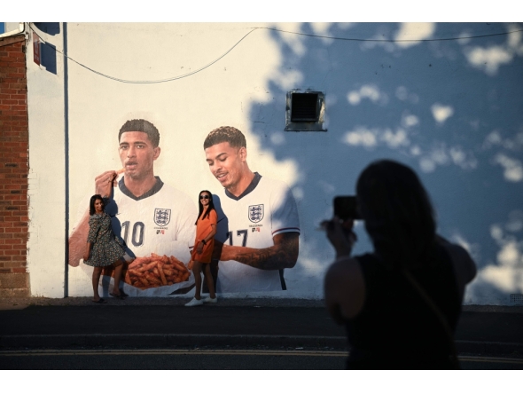People pose for a photo with a mural of England footballers Jude Bellingham and Morgan Rogers, eating a portion of chips, created by artist Dion Kitson, on the wall of a fish and chip shop near Halesowen in central England, on July 14, 2026. (Photo by Oli Scarff / AFP)