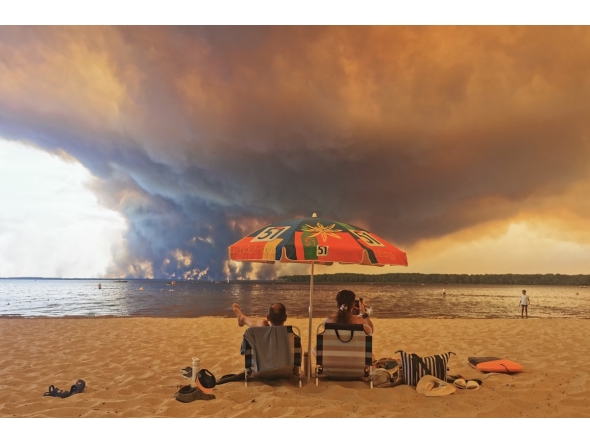 Beach goers sit on the beach of Moutchic in Lacanau, southwestern France on July 24, 2026, as clouds of smoke rise into the sky from a wildfire. Photo by Maximilien Lamy / AFP