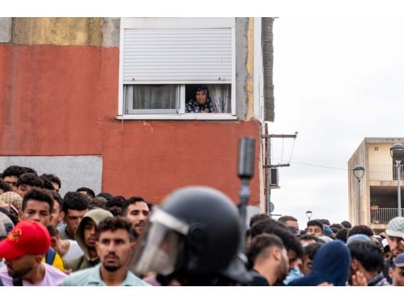 A resident watches from her window migrants waiting on the sidewalk before being registered in Loma Colmenar district of Ceuta on August 31, 2026. Photo by Antonio Sempere / AFP