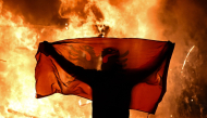 A protester holds an Albanian National flag in front of a burning kiosk during an anti-government demonstration in central Tirana on October 15, 2026, as protests continued for a third consecutive day over university conditions and demands for the resignation of Albanian Prime Minister. Photo by Armend NIMANI / AFP