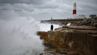 Members of the public stand on rocks as waves break near Portland Bill Lighthouse in Portland, southern England on October 23, 2026, as Storm Austen brings strong winds and heavy rain across much of southern England. (Photo by Henry Nicholls / AFP)