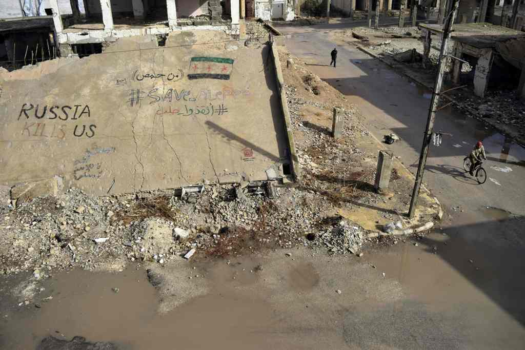 A Syrian rides a bicycle past a collapsed building on which graffiti in Arabic and English reads 