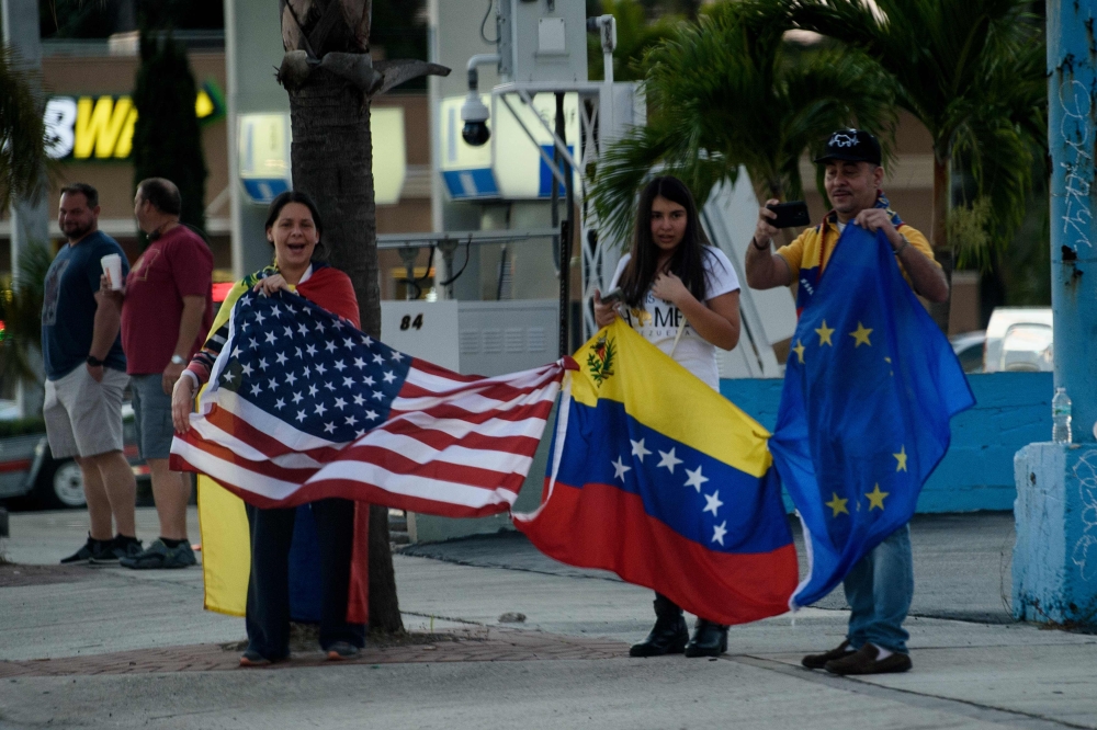 People holding the United States, Venezuelan and the European flag watch as a motorcade with US President Donald Trump travels from Mar-a-Lago to Trump International Golf Club for a Super Bowl party February 3, 2019 in West Palm Beach, Florida. / AFP / Br