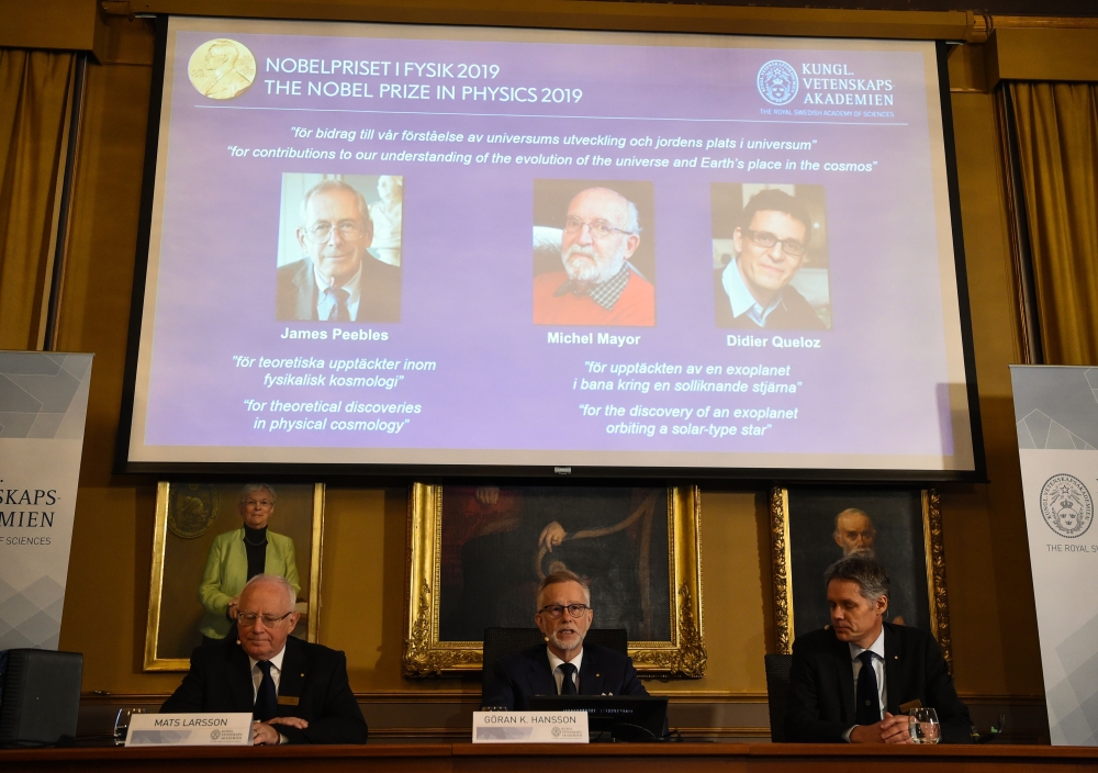Members of the Nobel Committee for Physics (Bottom L-R) Chair of the Nobel Committee Mats Larsson, Secretary General of the Academy Goran K Hansson, and Ulf Danielsson sit in front of a screen displaying the portraits of the winners of the 2019 Nobel Priz