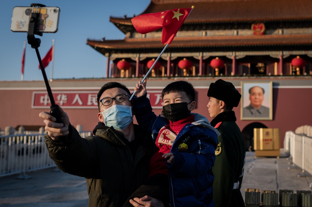  A boy waves a national flag as his dad holds him and uses a smartphone with a selfie stick to take a photo, both wearing protective masks, in front of the portrait of late communist leader Mao Zedong (R, back) at Tiananmen Gate in Beijing on January 23, 
