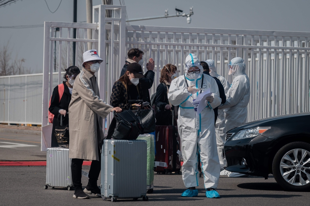 A worker (front R) wearing a protective suit as a preventive measure against the COVID-19 coronavirus stands next to passengers (L) at the New China International Exhibition Centre, near Beijing Capital Airport in Beijing on March 16, 2020./ AFP / NICOLAS