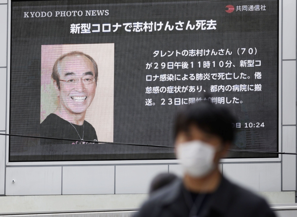 A man wearing a protective face mask, following an outbreak of the coronavirus disease (COVID-19), walks past in front of a huge screen reporting death of Japanese comedian Ken Shimura, who had been hospitalised after being infected with the new coronavir