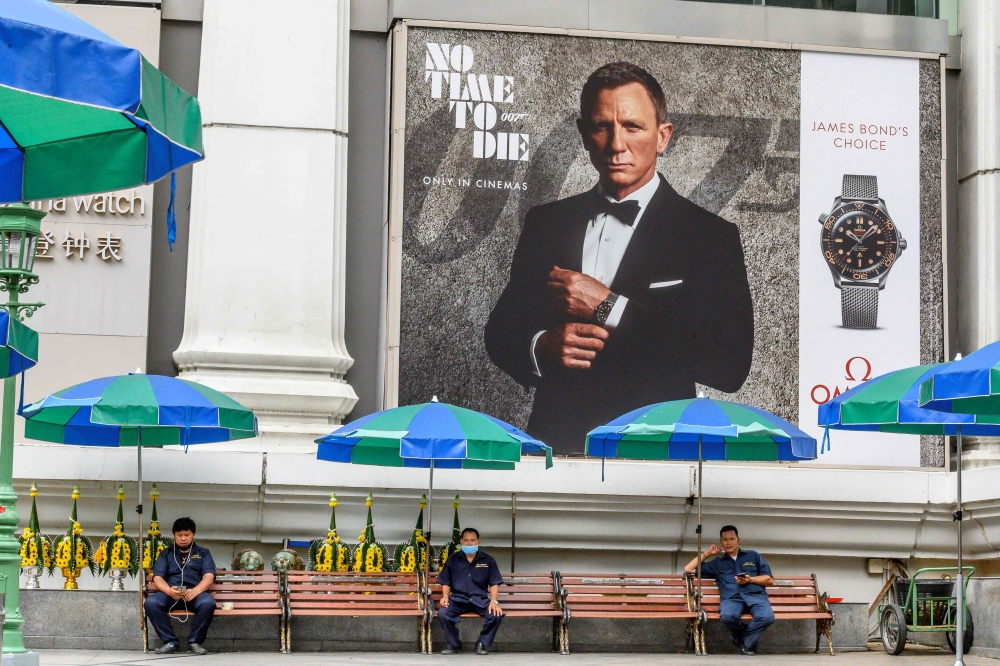 Security guards sit in the Erawan Shrine compound under an advertisement poster featuring Daniel Craig in the new James Bond movie 