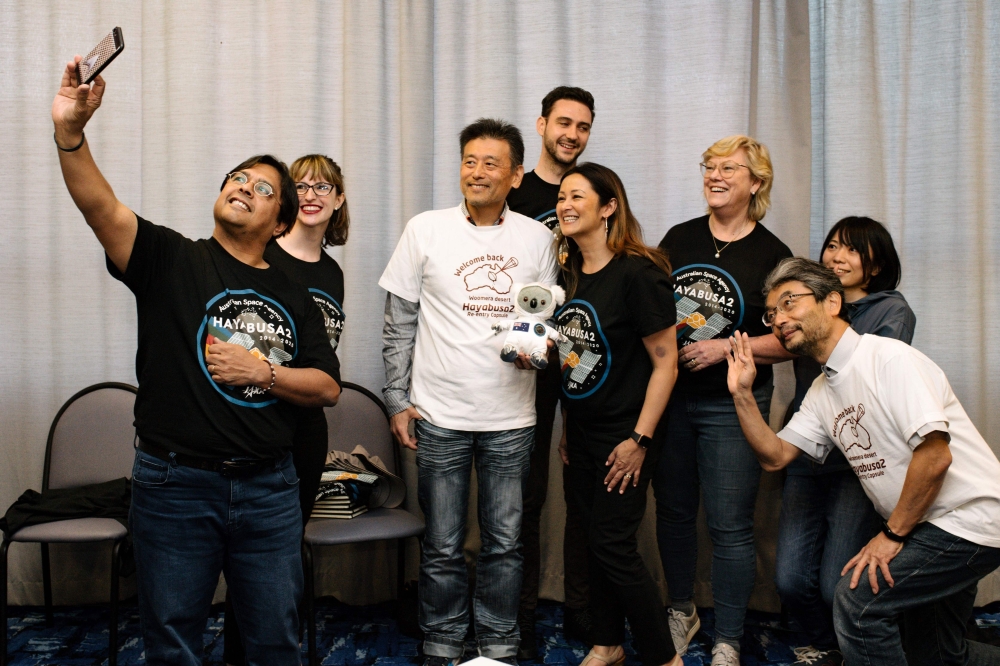 From the Japan Aerospace Exploration Agency (JAXA) and Australian Space Agency, (L-R) Karl Rodrigues, Caitlin Caruana, Masaki Fujimoto, Thomas Lewis, Jacqui Tyack, Megan Clarke, Nobuyoshi Fujimoto and Sakiko Kameruka pose for a selfie at a press conferenc