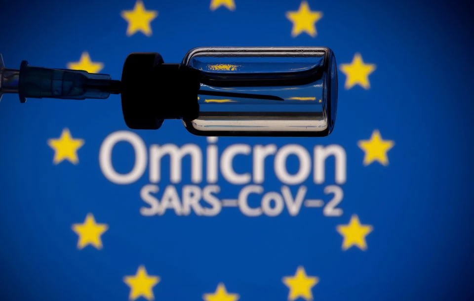 A vial and a syringe are seen in front of a displayed EU flag and words 