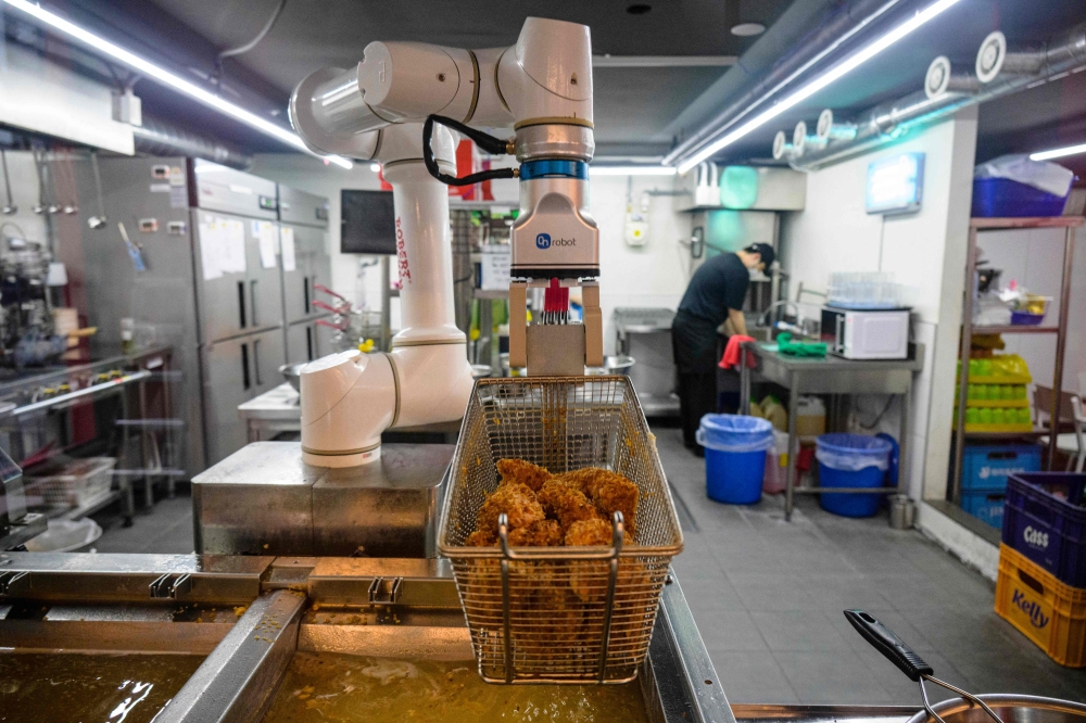 In this photo taken on June 13, 2023, a robot fries chicken as an employee washes dishes at a Robert Chicken restaurant in Seoul. (Photo by Anthony Wallace / AFP)