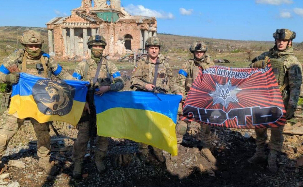 This handout photograph released by Ukrainian presidential chief of staff Andriy Yermak on September 17, 2023, shows Ukrainian servicemen posing for a photo with Ukrainian national flags in front of a destroyed building in the village of Klyshchiivka, Donetsk region. (Photo by Handout / Ukrainian Presidential Chief of Staff Andriy Yermak / AFP)