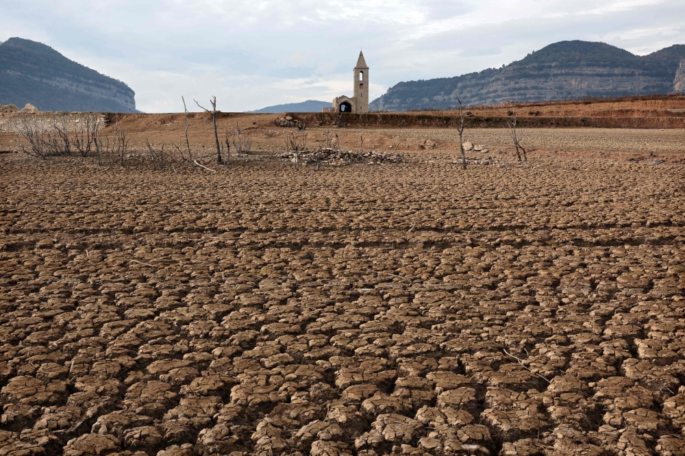 This photograph taken on January 15, 2024 shows the dry soil next to the low water-level reservoir of Sau with in background Sant Roma de Sau church, in the province of Girona in Catalonia. Catalonia struggles with historic drought for three years, with some residents already experiencing water restrictions in their daily life. (Photo by LLUIS GENE / AFP)