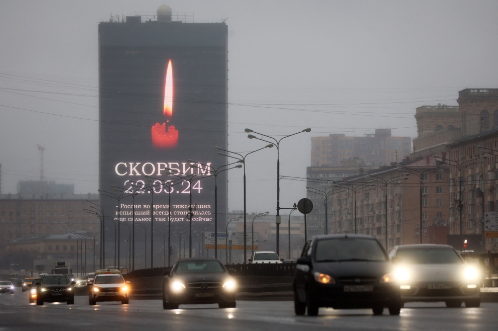 Cars drive past an advertising screen on the facade of a building displaying an image of a lit candle and the slogan 