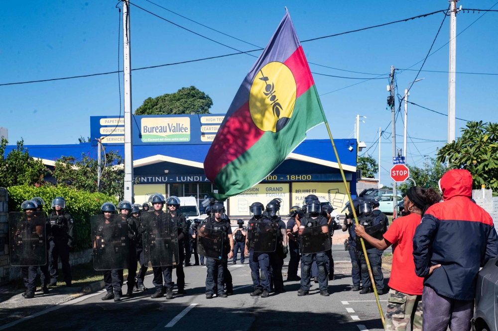 A pro-independence supporter holds a Kanak and Socialist National Liberation Front (FLNKS) flag in front of police officers outside the headquarters of the Union Caledonienne (UC), after police intervened in the UC offices this morning, where a press conference of the Cellule de Coordination des Actions de Terrain (CCAT) was due to be held, in Noumea, on June 19, 2024. Photo by Delphine Mayeur / AFP.

