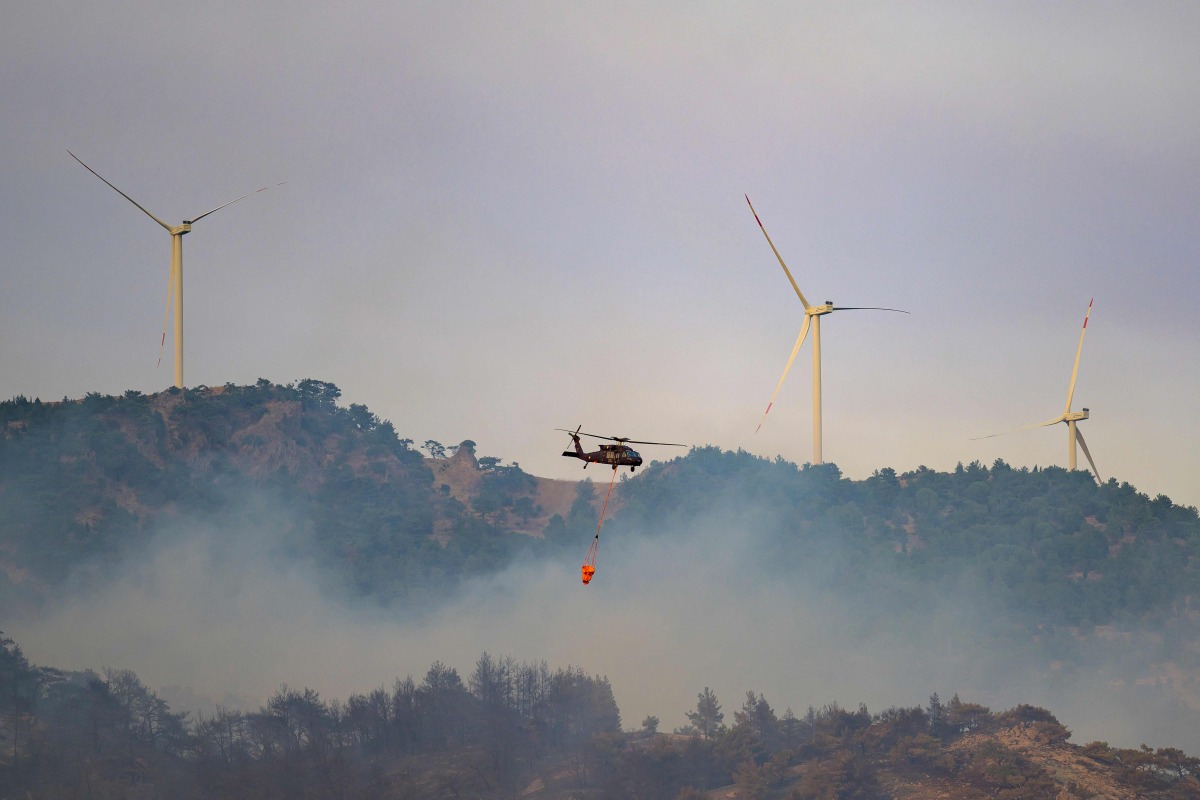 This photograph shows a helicopter carrying water to fight a forest fire, with windmills in the background in Turkey's western province of Izmir on August 17, 2024. (Photo by Yasin AKGUL / AFP)