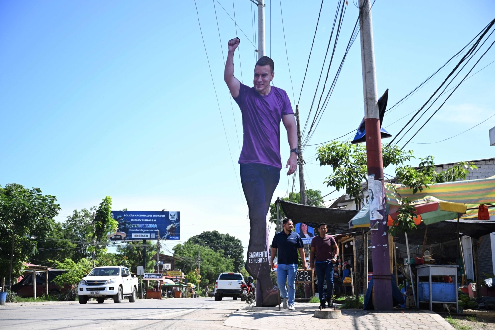People walk past a giant cardboard image of Ecuador's President Daniel Noboa in Olon, Santa Elena province, Ecuador, on April 14, 2025, the day after the presidential runoff election.  (Photo by Raul Arboleda / AFP)