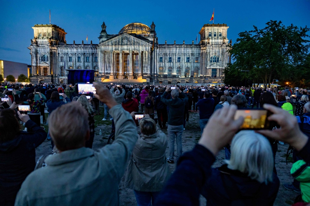 People watch the Reichstag building illuminated in a tribute to the artwork of artists Christo and Jeanne-Claude, in Berlin on June 9, 2025. (Photo by Odd Andersen / AFP)