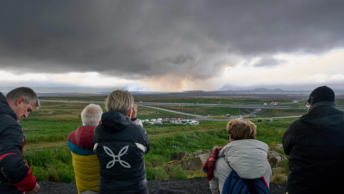 Tourists standing next to a memorial in Keflavik, Iceland on July 16, 2025, observe from a distance smoke and lava emanating from a volcano near Grindavik on the Icelandic peninsula of Reykjanes following its eruption. Photo by Halldor KOLBEINS / AFP