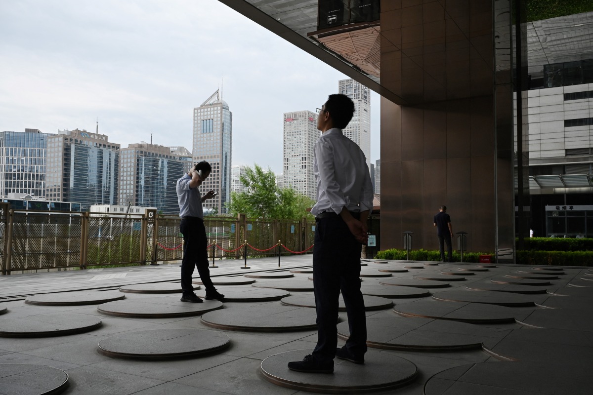Men relax under an office building in Beijing’s central business district on July 31, 2025. (Photo by GREG BAKER / AFP)