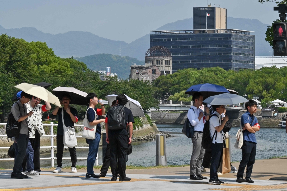 A group of tourists shelter under umbrellas in the summer heat as they wait for the light to change, with the Atomic Bomb Dome in the background in downtown Hiroshima on August 5, 2025. (Photo by Richard A. Brooks / AFP)