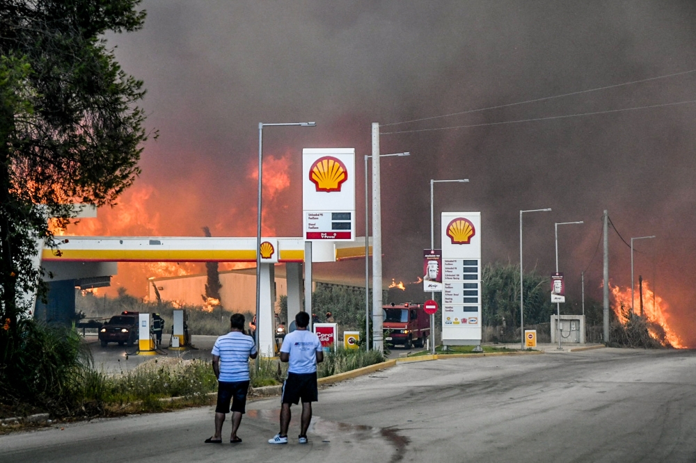 Inhabitants stand on a road as a wildfire approaches a gas station near the city of Patras, western Greece on August 12, 2025. (Photo by Eurokinissi / AFP) 