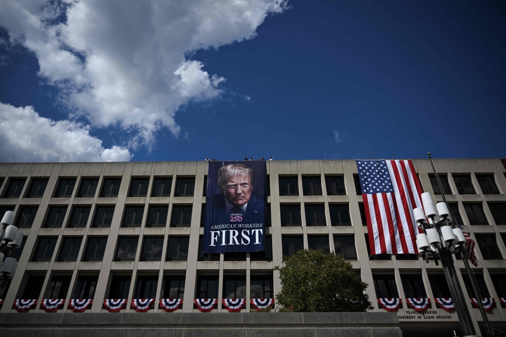 Workers hang a large photo of US President Donald Trump next to a US flag on the facade of the Department of Labor headquarters building in Washington, DC, on August 27, 2025. (Photo by Drew Angerer / AFP)