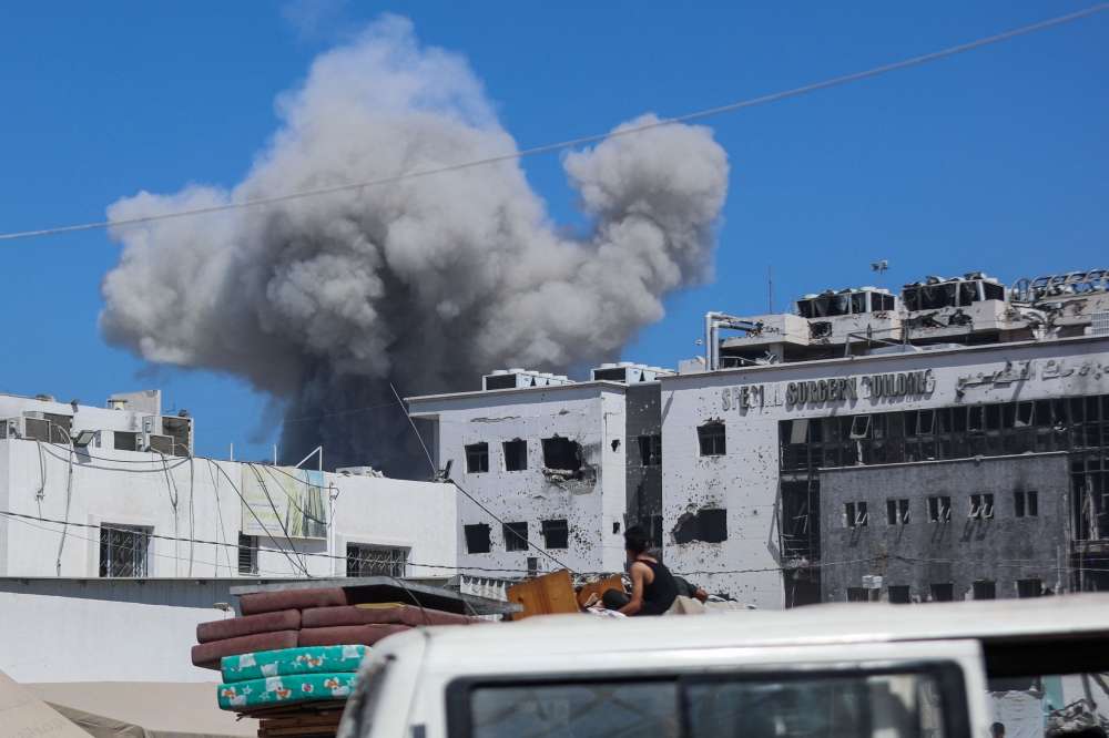 Displaced Palestinians sitting in the back of a truck laden with their belongings while fleeing bombardment, watch smoke billowing amid Israeli strikes on Al-Shati camp for Palestinian refugees west of Gaza City on September 18, 2025. (Photo by Omar Al-Qattaa / AFP)
