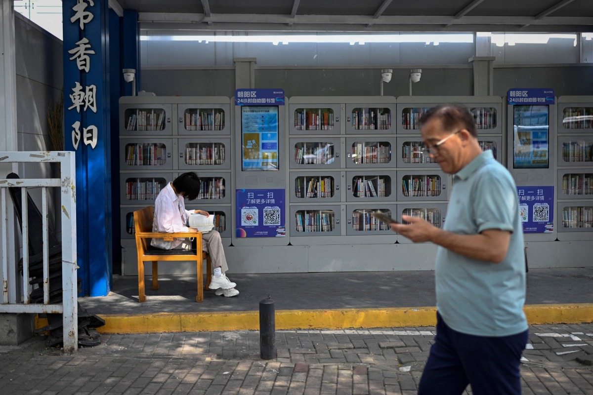A woman (L) reads a book at a self-service library beside a pavement in Beijing on September 22, 2025. (Photo by WANG Zhao / AFP)