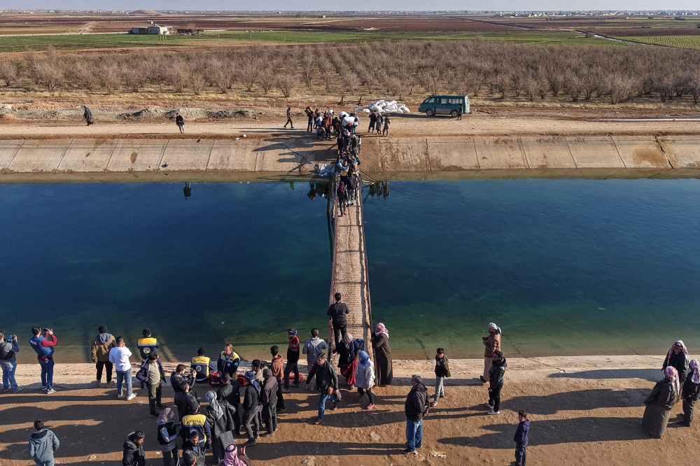 This aerial photograph shows people along with their belongings walking across a damaged bridge as they flee from a Kurdish-controlled area, in Rasm al-Harmal, east of Aleppo city on January 16, 2026. (Photo by Bakr ALkasem / AFP)