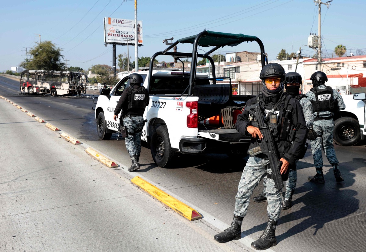 National Guard members stand guard near a burnt bus set on fire by organised crime groups in response to an operation in Jalisco to arrest a high-priority security target, at one of the main avenues in Zapopan, state of Jalisco, Mexico, on February 22, 2026. (Photo by Ulises RUIZ / AFP)