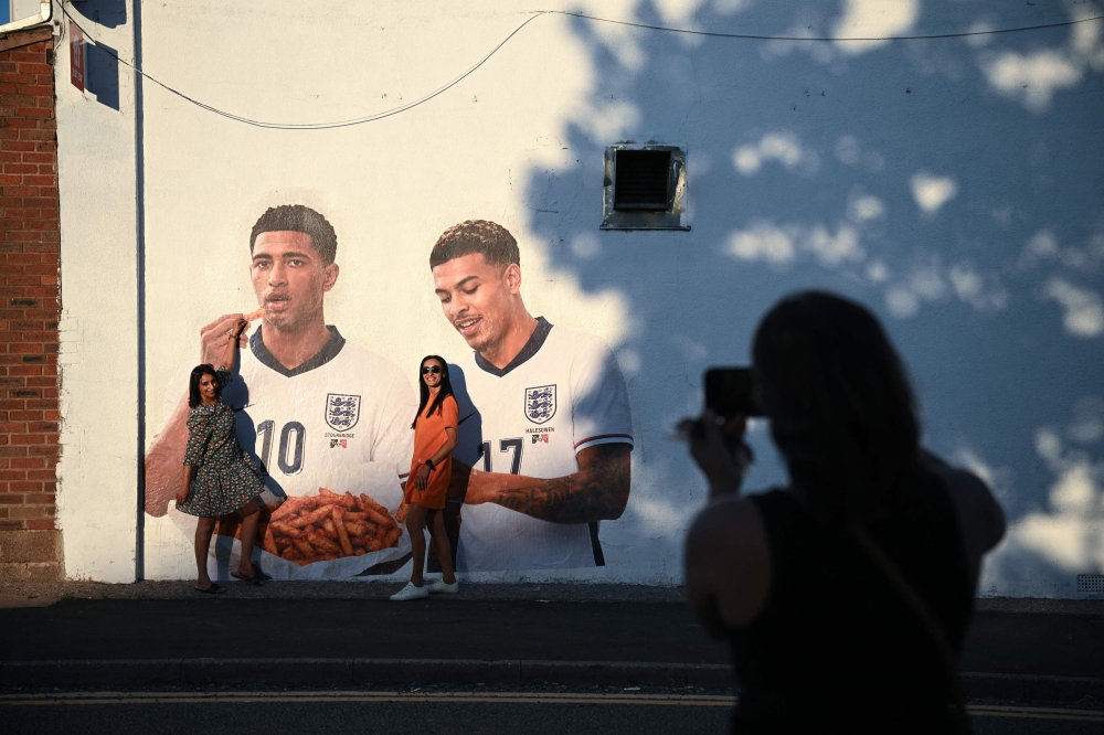 People pose for a photo with a mural of England footballers Jude Bellingham and Morgan Rogers, eating a portion of chips, created by artist Dion Kitson, on the wall of a fish and chip shop near Halesowen in central England, on July 14, 2026. (Photo by Oli Scarff / AFP)