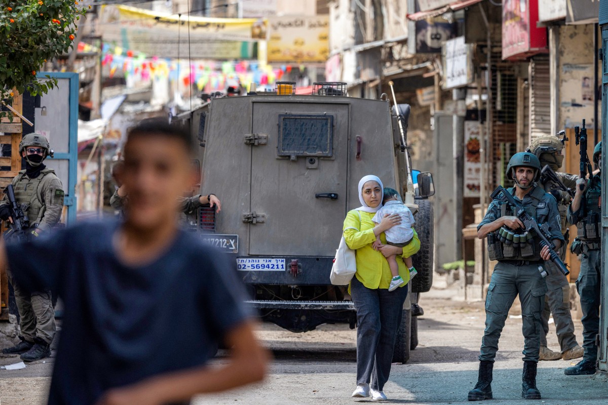 A Palestinian woman carrying a child walks past members of the Israeli occupation army and armoured vehicles during an Israeli military operation at the Balata camp for Palestinian refugees, east of Nablus in the occupied West Bank, on August 31, 2026. Photo by JAAFAR ASHTIYEH / AFP