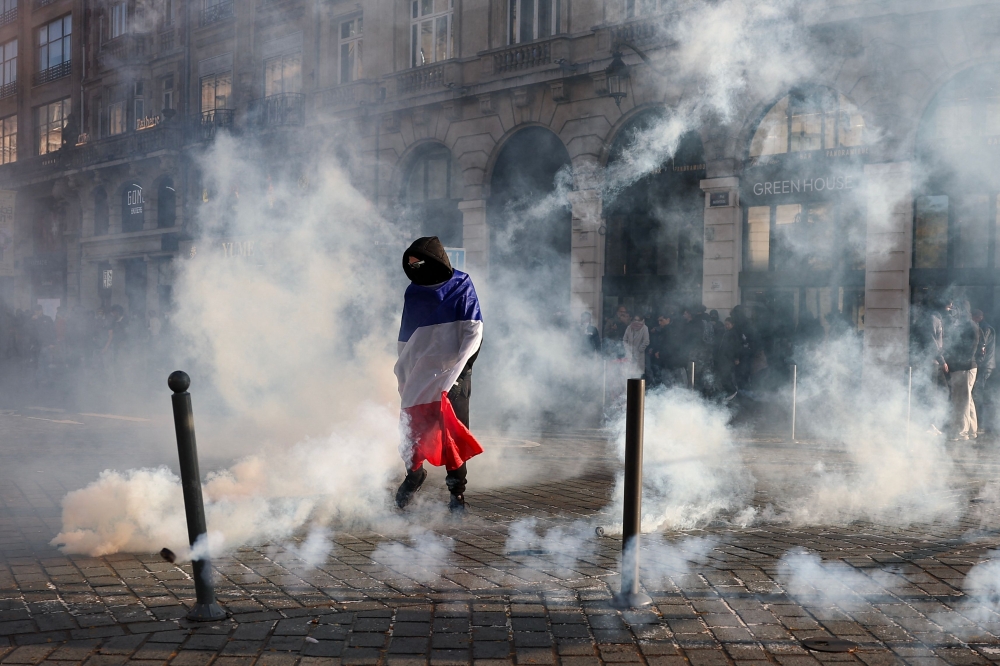 A protester, wrapped in a flag of France, stands in the smoke at Place de l'Opera during ongoing protest action, in Lille, northern France, on October 17, 2026. (Photo by Sameer Al-Doumy / AFP)
