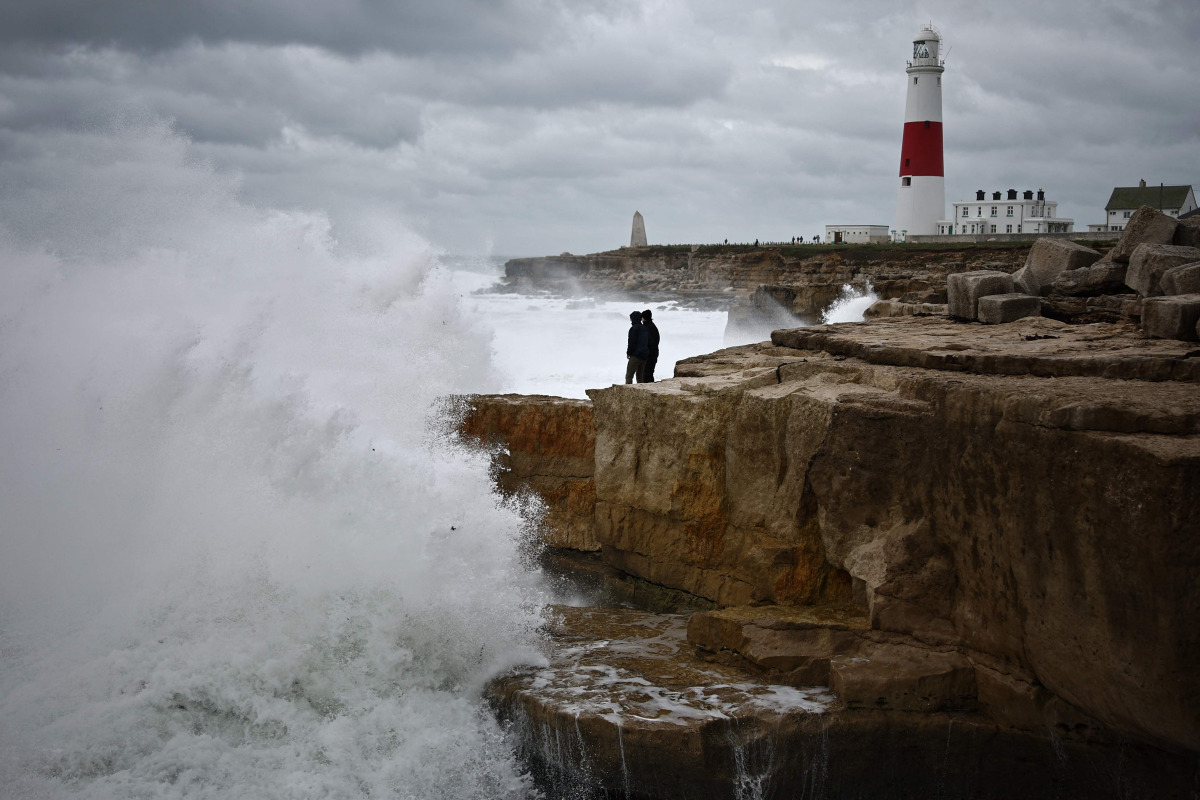 Members of the public stand on rocks as waves break near Portland Bill Lighthouse in Portland, southern England on October 23, 2026, as Storm Austen brings strong winds and heavy rain across much of southern England. (Photo by Henry Nicholls / AFP)